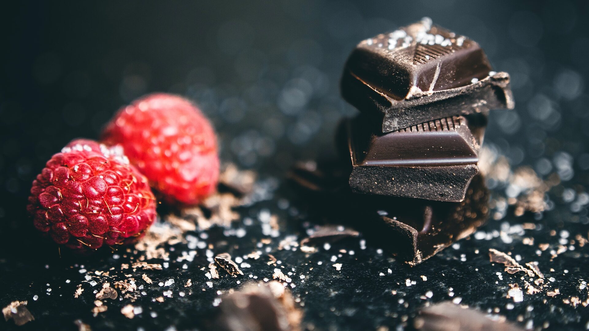 Een donkere achtergrond met op de voorgrond een stapeltje met blokjes chocolade en een paar frambozen