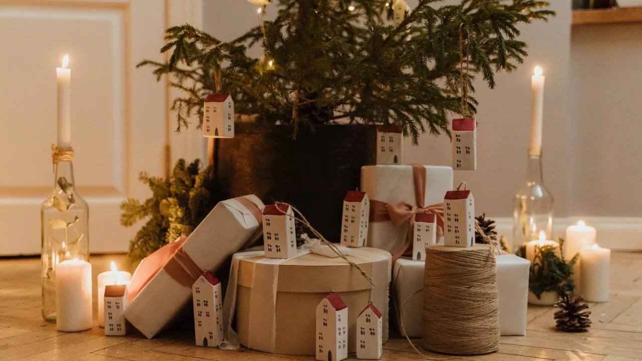 een kerstboom in een donkerbruine pot met een stapel cadeautjes eronder. De cadeautjes zijn doosjes in verschillend vormen en maten en ze zijn allemaal wit en verschillende tinten beige. Er staan kleine huisjes tussen de cadeautjes.