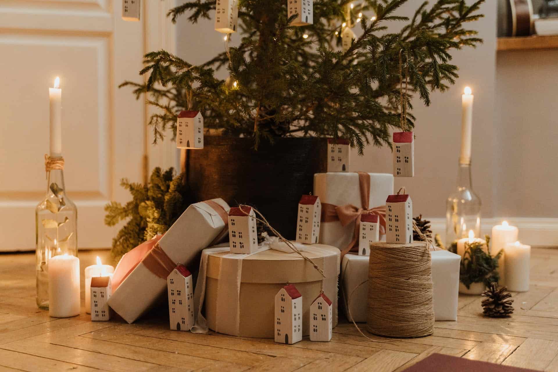 een kerstboom in een donkerbruine pot met een stapel cadeautjes eronder. De cadeautjes zijn doosjes in verschillend vormen en maten en ze zijn allemaal wit en verschillende tinten beige. Er staan kleine huisjes tussen de cadeautjes.