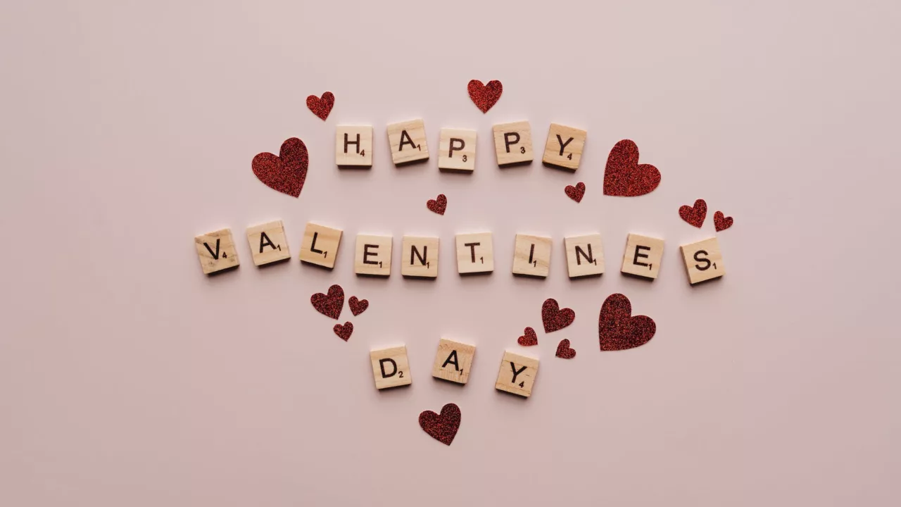 Een roze achtergrond met rode hartjes en scrabble letters die samen "Happy Valentine's Day" spellen.