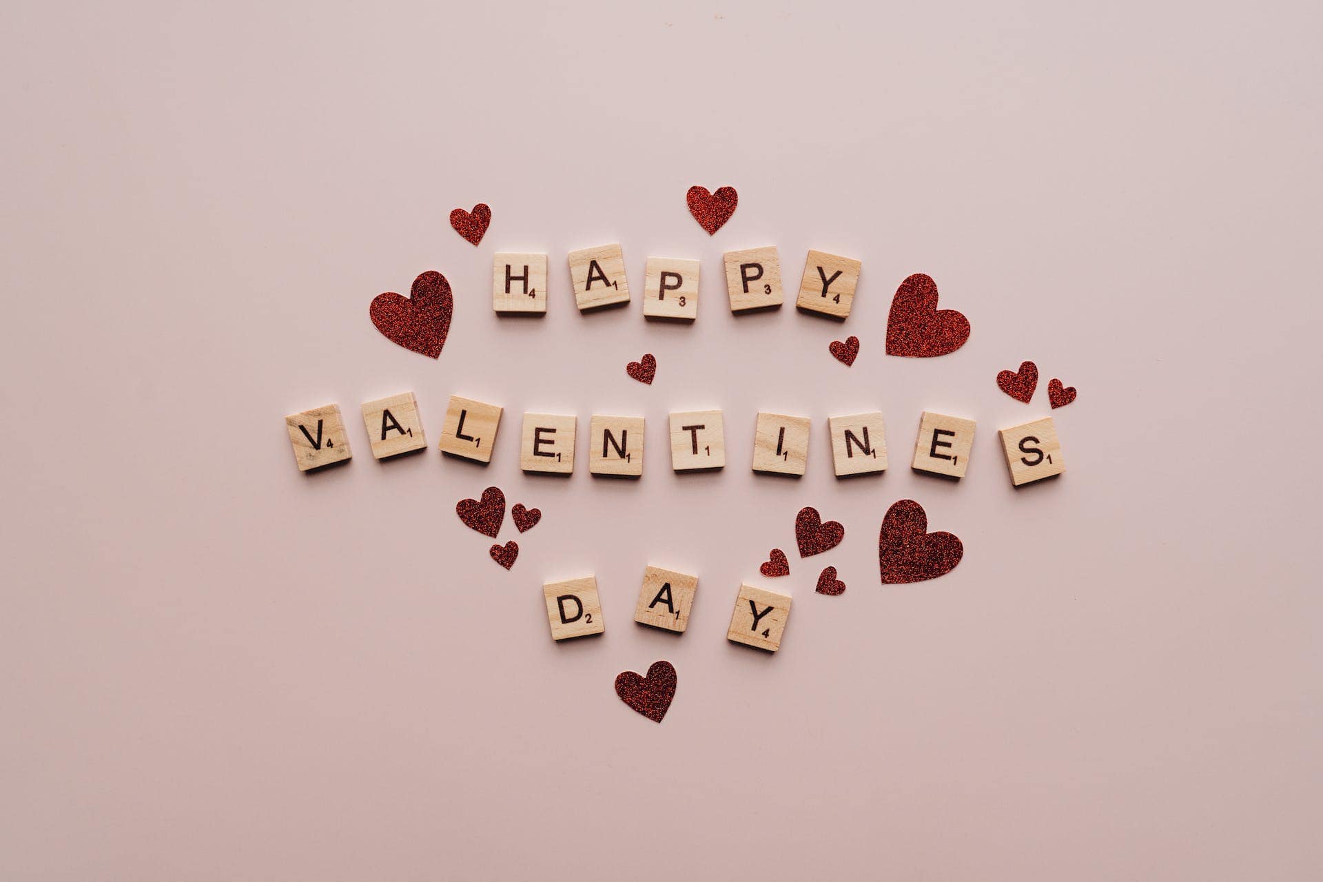 Een roze achtergrond met rode hartjes en scrabble letters die samen "Happy Valentine's Day" spellen.